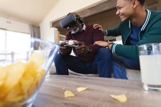 Happy african american father using vr headset and gamepad at home with adult son