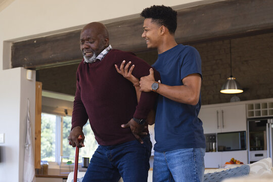 Happy african american adult son helping senior father stand up from couch at home