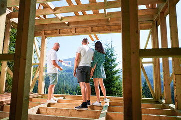 Man designer with plan showing to couple the process of constructing wooden framed house. Back view...