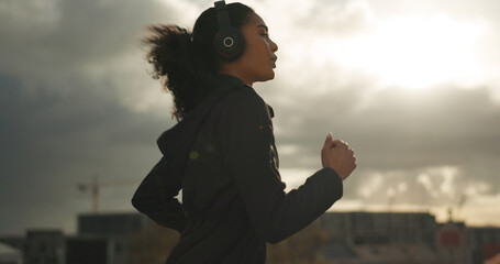 Exercise, music and a sports woman running in the city for health or cardio preparation of a marathon. Fitness, wellness or training and a young runner or athlete listening to audio with headphones - obrazy, fototapety, plakaty