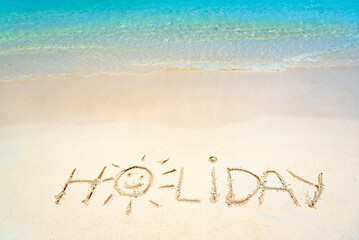 Holiday hand written in the sand on the beach blue waves in the background. Sun drawing on golden...
