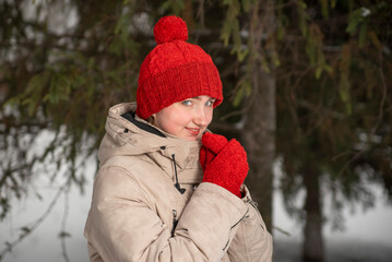 Fototapeta na wymiar Beautiful young woman with a sly look in knitted red hats and mittens. Winter forest or park background. Winter walk