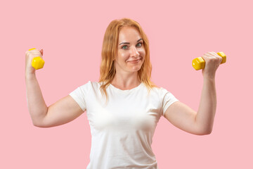 Powerful young woman showing muscles on arms with dumbbells. Portrait of lovely red hair girl on pink pastel background