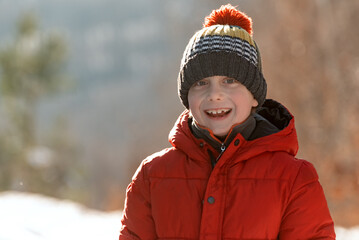 Funny smiling Caucasian boy in blown red jacket and hat with bubo in winter outside. Portrait of happy boy