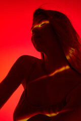 Sensual, tender young woman in top posing with eyes clothe on red background with neon reflection...
