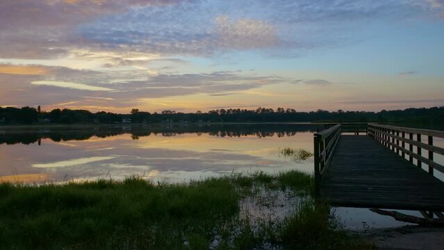 In this captivating timelapse captured in Ocoee, Florida. Clouds glide over a tranquil lake. Subtle shifts in the sky's hue paint a mesmerizing canvas. A quaint wooden pier extends into the water.