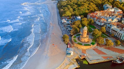 Drone photo captures Kołobrzeg's maritime charm, featuring the iconic lighthouse, cerulean sea, turbulent waves, a distant pier, and autumnal hues on the trees. - obrazy, fototapety, plakaty
