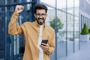 Joyful successful man looking at camera, businessman holding phone in hands, celebrating victory...