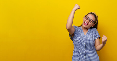 Excited satisfied young Asian woman wearing a dress checkered with glasses raising her fists up,...