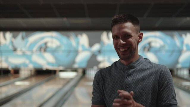 Middle aged caucasian man with hard shadows on his face smiles in bowling club, applauds and shows thumbs up. Happy player in front of bowling alley under light source after successful throw.