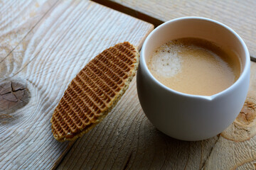 A white cup of black coffee with a cookie is standing on the wooden table. A cup of coffee with a brown biscuit on the wooden background
