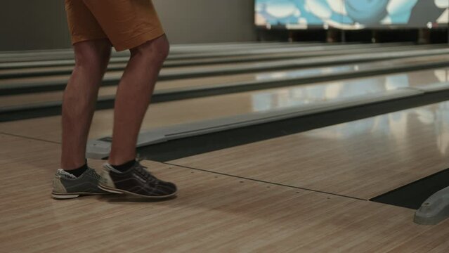 Man wearing rental bowling shoes throws ball down varnished wooden lane at public bowling alley. Close up of bowlers legs in dark club and attempting shots at pins.