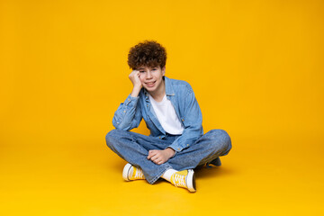 Curly boy teenager sitting on the floor, on a yellow background.