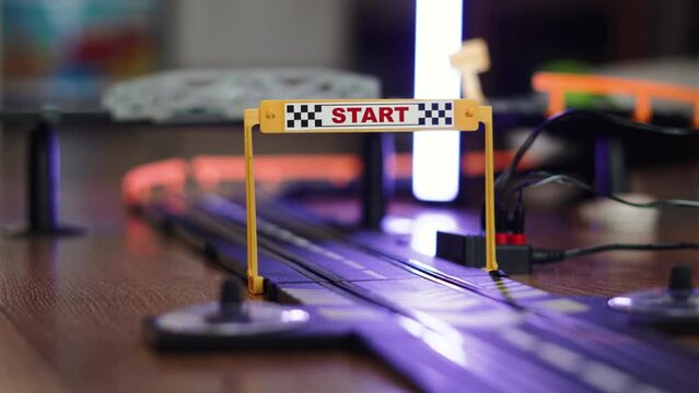 Toy electric slot cars drive around track through Start frame with checkered flag after loop. Formula cars race along slots with conductive rails during home play, no people. Sports rivalry in game.
