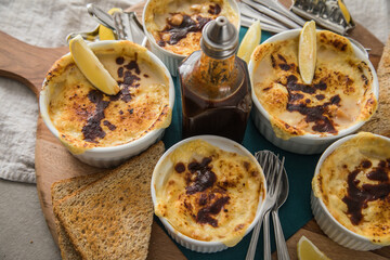 Typisches DDR Würzfleisch Ragout Fin mit Käse überbacken, Zitrone, Toast Brot und Worcester Sauce  in Auflaufform Schale auf Holz Brett, Tuch und hell grau Stein Hintergrund
