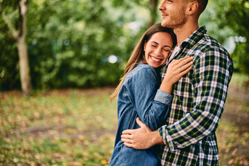 A lovely couple looking happy, a girl in her boyfriends hug, enjoying the nature during the autumn season. - obrazy, fototapety, plakaty