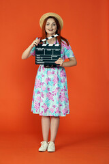 Woman in hat and dress holding clapper board, looking and smiling at the camera, isolated on orange background. Portrait of female traveler.