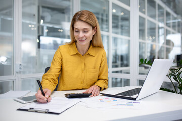 Successful mature female financial accountant at workplace inside office, fills data in table,...