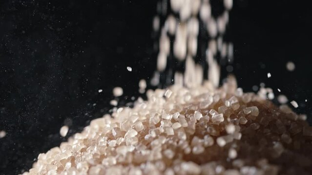 Brown cane sugar is poured into pile. Macro shot slow motion black background. Minimizing sugar consumption is an effective strategy to prevent obesity, diabetes, and other grave conditions. Nutrition