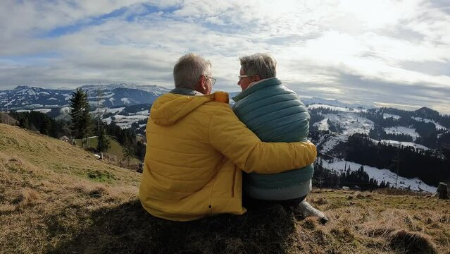 Back view of senior couple hugging each other while resting and enjoy landscape at mountain peak during hiking winter day - Travel concept