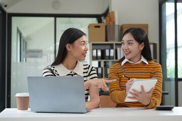 Two diverse mixed race beautiful african and asian business women wearing casual clothes, smiling with happiness, talking while taking break in indoor office, looking at laptop, holding cup of coffee