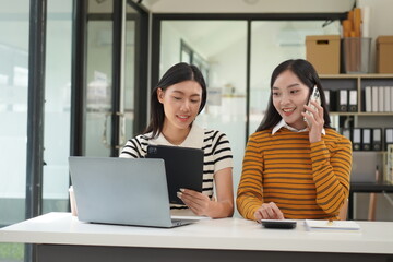 Two diverse mixed race beautiful african and asian business women wearing casual clothes, smiling with happiness, talking while taking break in indoor office, looking at laptop, holding cup of coffee