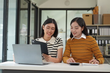 Two diverse mixed race beautiful african and asian business women wearing casual clothes, smiling with happiness, talking while taking break in indoor office, looking at laptop, holding cup of coffee
