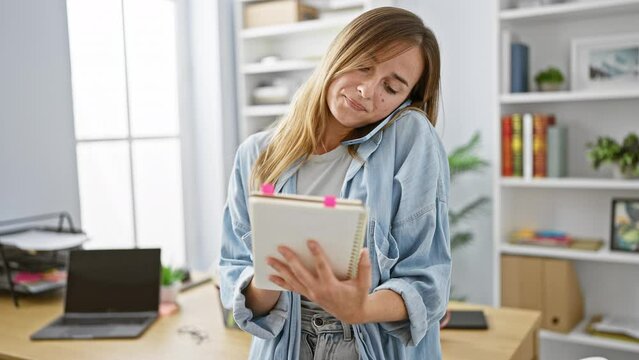 Bright-eyed young blonde woman, a confident business worker engrossed in a captivating talk over her smartphone at the office desk, diligently taking key notes.
