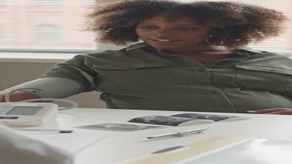 Vertical medium shot of young pregnant Black woman having her blood pressure checked during medical appointment at modern doctor office