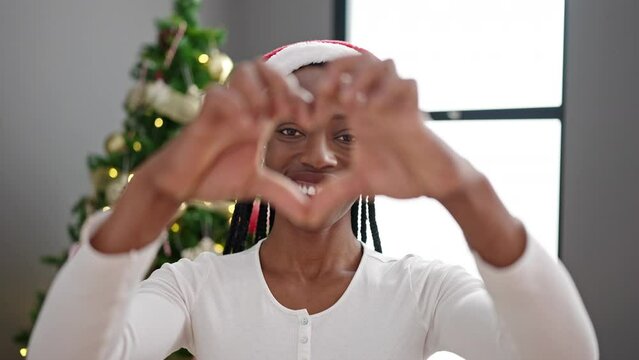 African american woman doing heart gesture standing by christmas tree at home