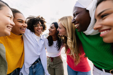 Seven happy young adult women from different cultures laughing together outdoors. Female friendship concept with diverse group of girls friends hugging each other having fun at city street