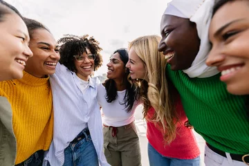 Fotobehang Seven happy young adult women from different cultures laughing together outdoors. Female friendship concept with diverse group of girls friends hugging each other having fun at city street © Xavier Lorenzo
