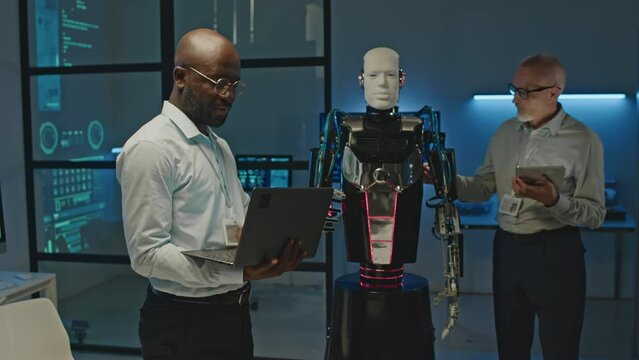 Medium shot of two multiethnic male robotics engineers discussing activation of humanoid robot while working together in scientific lab