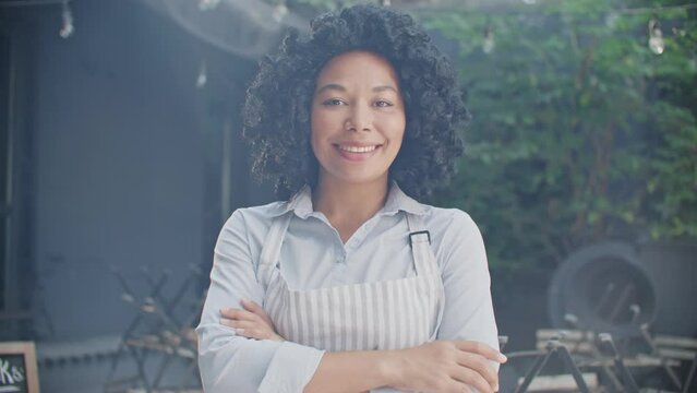Zooming in on African American waitress wearing apron while crossing hands at camera. Young female worker smiling with joy while standing on cafe terrace. Business. Serving concept.
