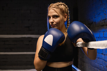 Strong female fighter in blue boxing gloves is training in the ring against a blue light background