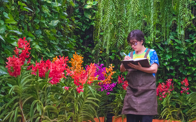 Asian adult woman in casual style is taking notes while studying the cultivation and propagation of colorful orchids in ornamental garden