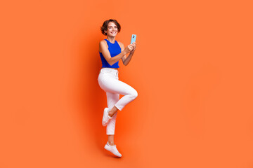 Full size profile portrait of pretty excited girl jumping use smart phone empty space isolated on orange color background