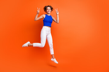 Full size portrait of energetic excited girl jumping demonstrate v-sign empty space isolated on orange color background