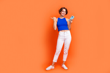 Full body portrait of gorgeous nice lady hold smart phone indicate finger empty space ad isolated on orange color background