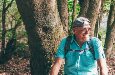 Naklejka na ściany i meble Portrait of smiling senior man with sunglasses and backpack sitting in a trekking day in the forest enjoying nature and healthy lifestyle in vacation or retirement