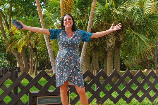 Woman standing smiling in a tropical park in Almunecar (Granada) with open arms and mobile phone in her hand in summer in a flowery dress.