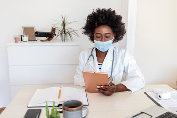 Female African American healthcare worker using digital tablet at her office in hospital. Wearing a face mask.