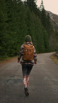Back view of the young woman traveller walking in middle of empty road in mountains, carrying backpack, enjoying of the nature