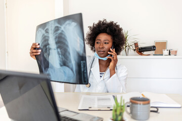 Doctor looking X-rays while having phone call. Young African-American doctor at office desk looking at an x-ray and talking on the phone. - obrazy, fototapety, plakaty