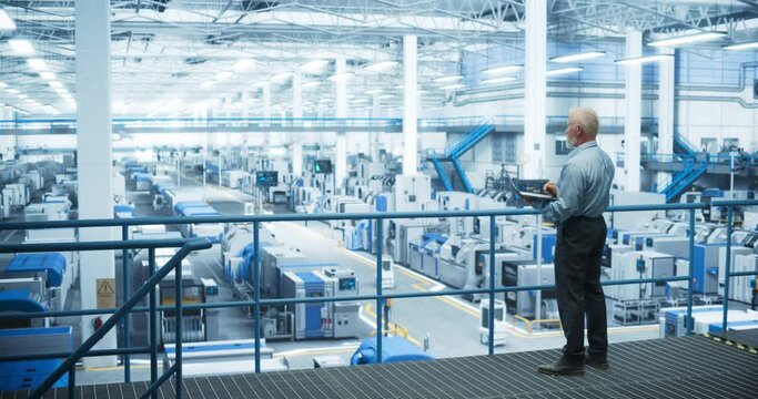 Senior Engineer Standing on a Platform, Using Laptop Computer at an Electronics Factory. Machines are Undergoing Maintenance, Specialist Monitoring the Progress Through Online Software. Static Footage