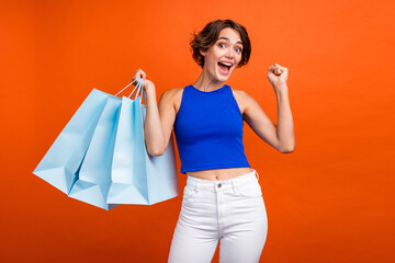 Portrait of delighted nice lady hold store mall bags raise fist success isolated on vivid orange color background