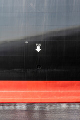 Section of the black and red hull of a ship with letters TUG and arrow pointing to waterline and location to secure tow of tugboat