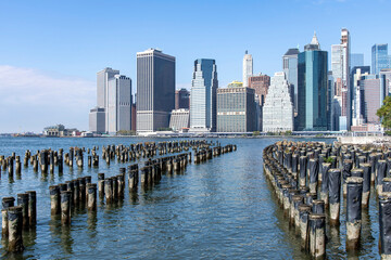 Hundreds of old wooden pier supports in the East River with panoramic view on skyscrapers and landmarks of Lower Manhattan, New York City, NY, USA against a clear blue sky - obrazy, fototapety, plakaty