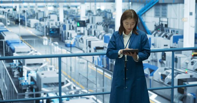 Thoughtful Asian Engineer Monitoring and Analyzing Conditions at a Pristine Modern Electronics Factory with Automated Robot Hands Working with the Help of Artificial Intelligence Software