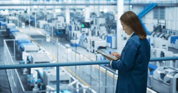 Young Asian Female Engineer Standing on a Platform, Using Tablet Computer at an Electronics Factory. Machines are Undergoing Maintenance, Specialist Monitoring the Progress Through Online Software